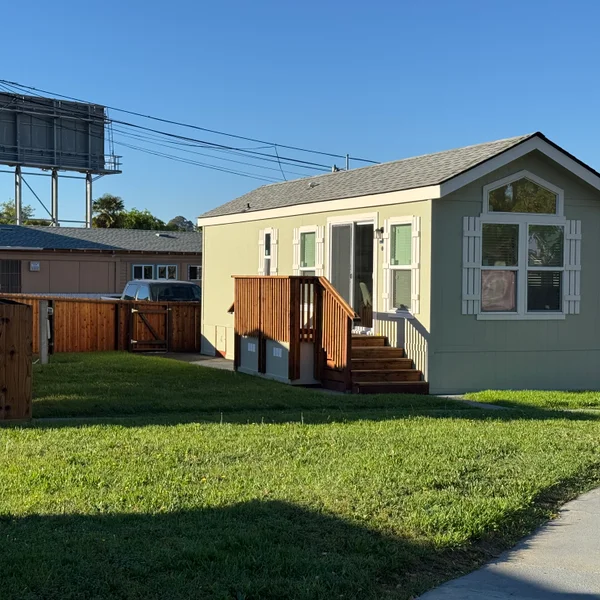 Light green manufactured home with white shutters features a small wooden deck and grassy lawn.