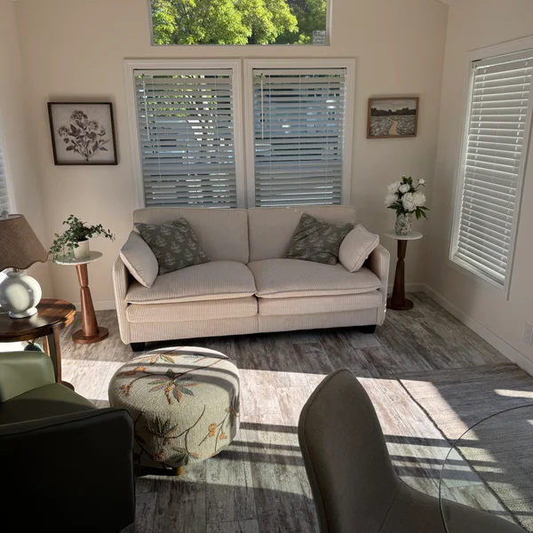 Bright real estate living room features a beige couch with floral pillows, accent chairs and ottoman, wood floors and blinds.