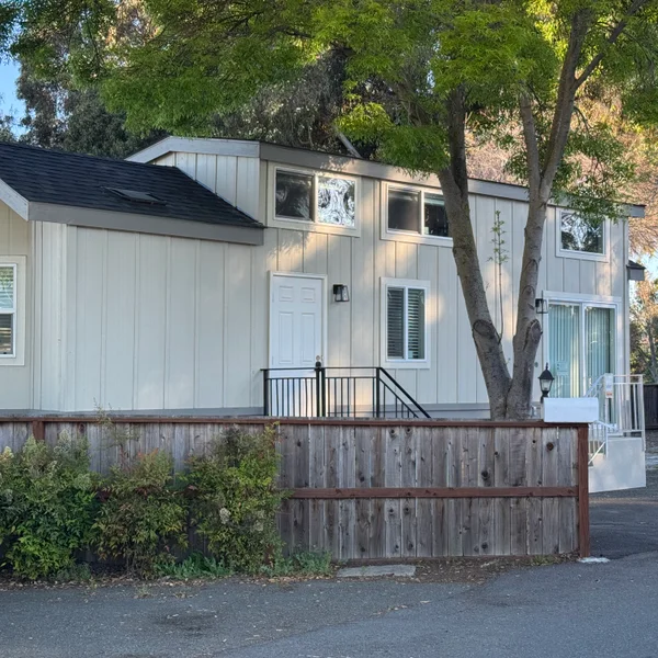 Real estate view shows a two-story, light-colored house with a wooden fence and mature tree.
